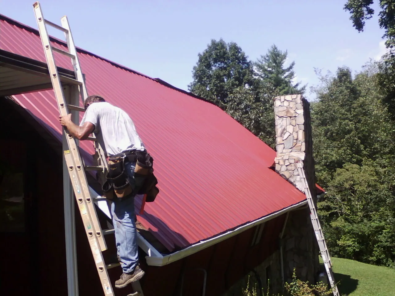 Roofer on a ladder beside a red metal roof and stone chimney — Soffit & Fascia Repair by Roofing Brothers in East Greenbush